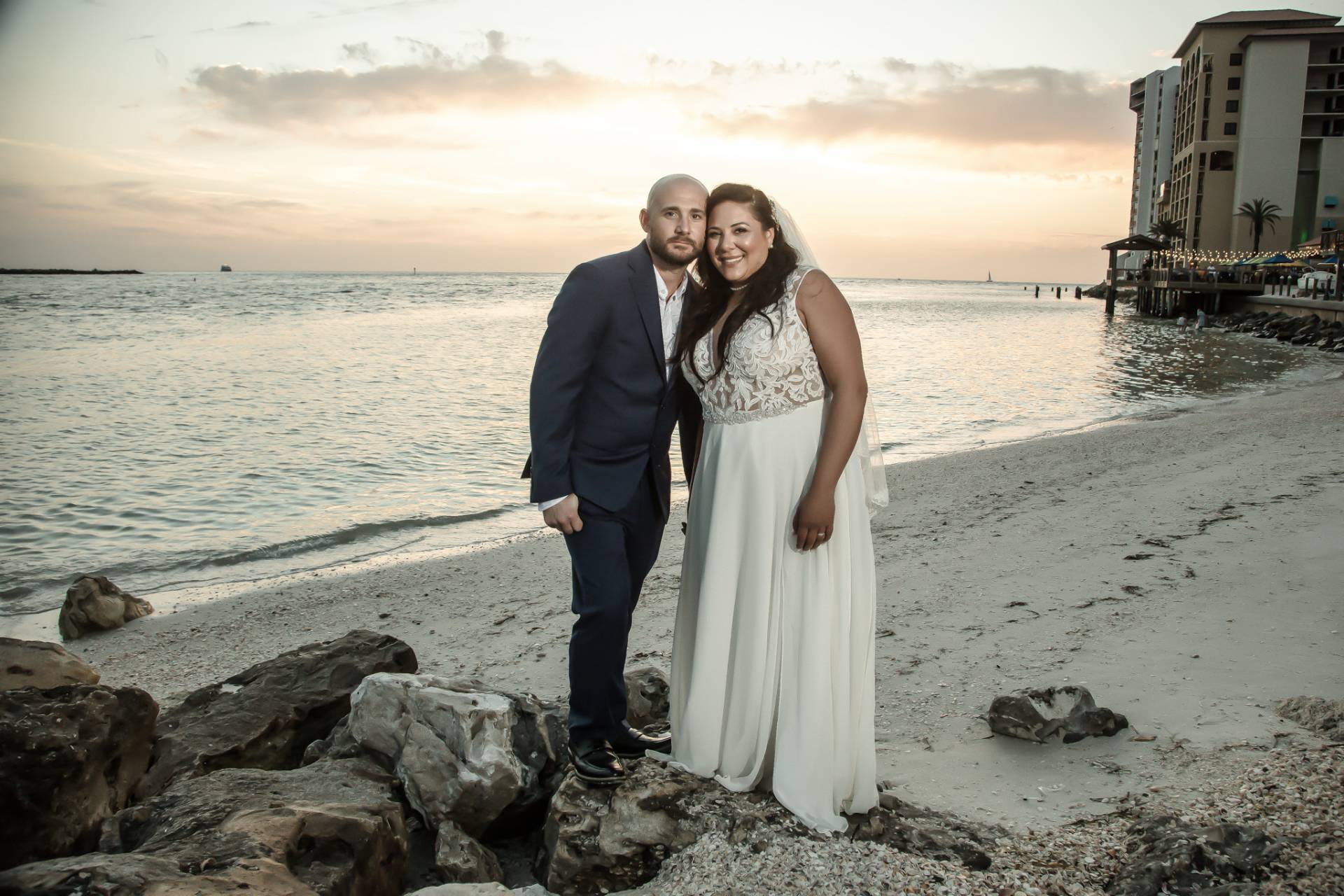 bride and groom standing on the beach at sunset near some rocks